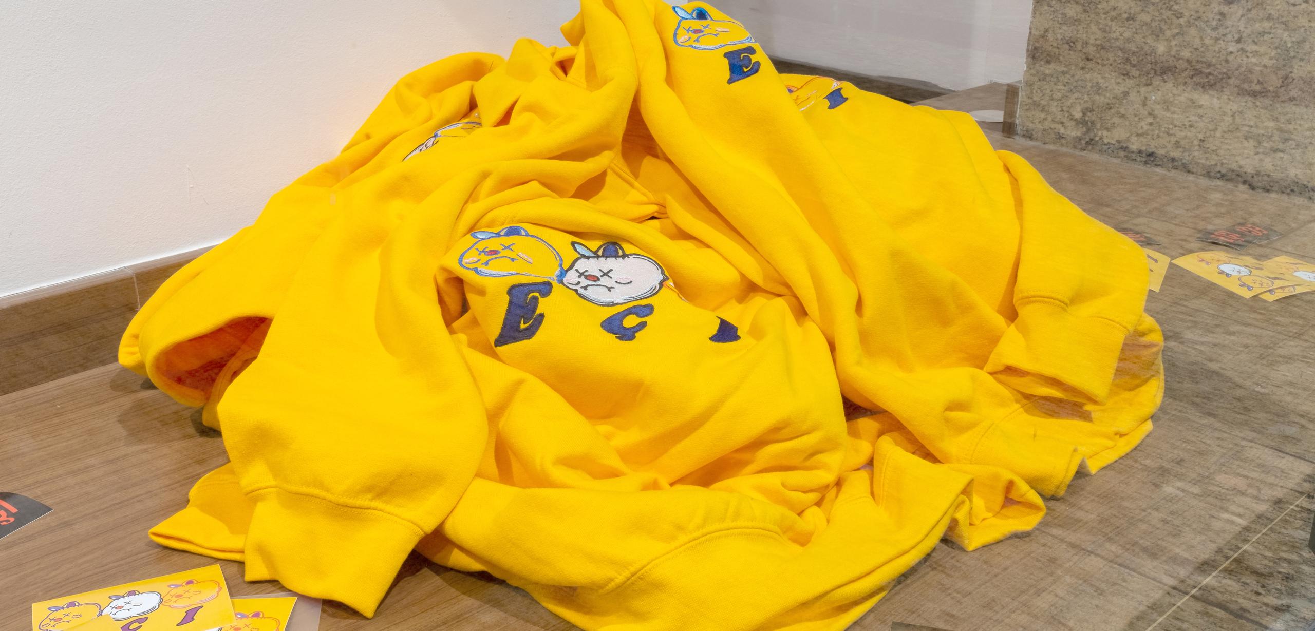 A pile of yellow hoodies with illustrated cat faces and "E" letters, laid on a wooden surface next to small printed cards.