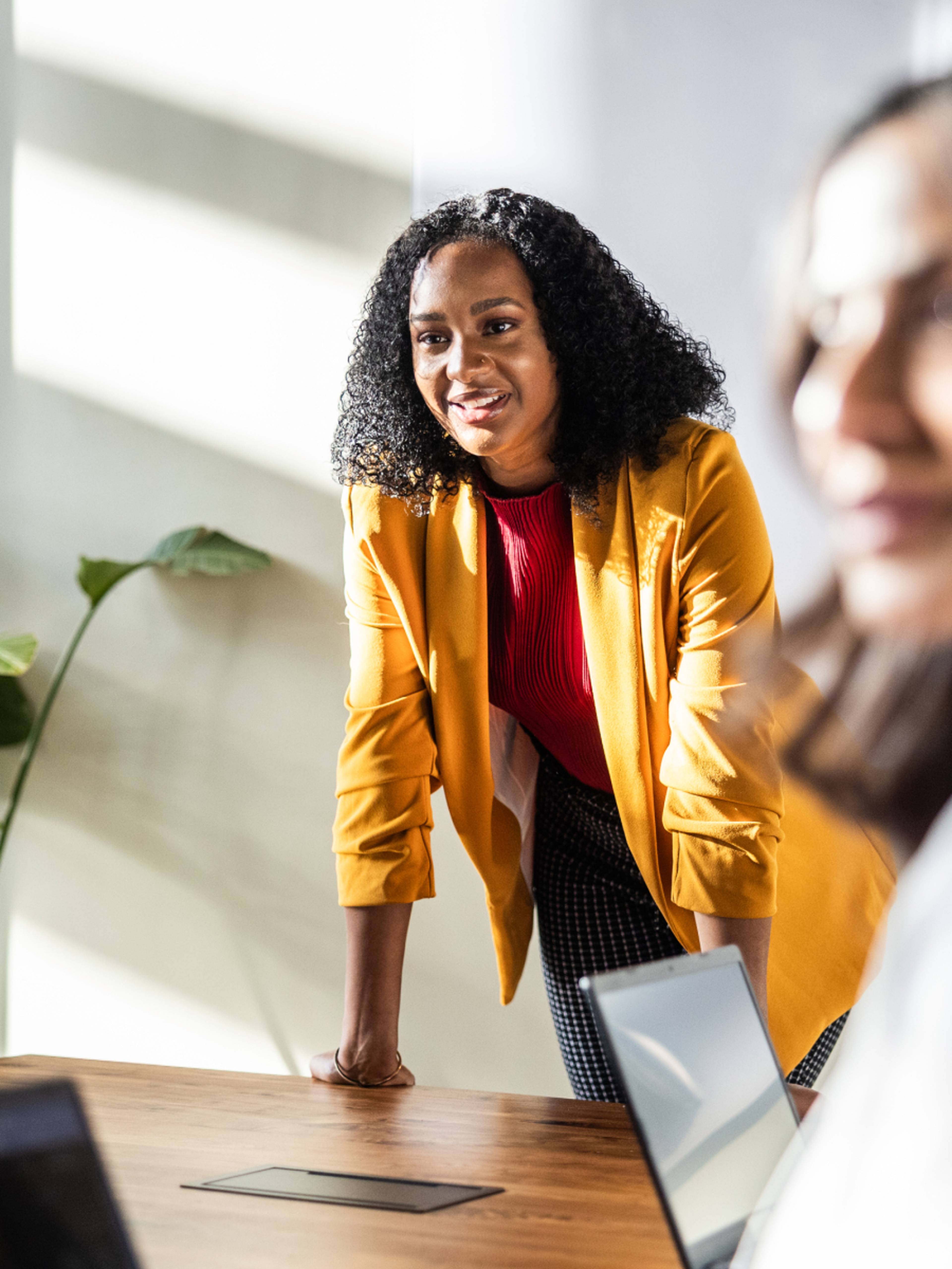Woman in a yellow blazer stands confidently at a desk, smiling during a meeting, with laptops and plants in the background.