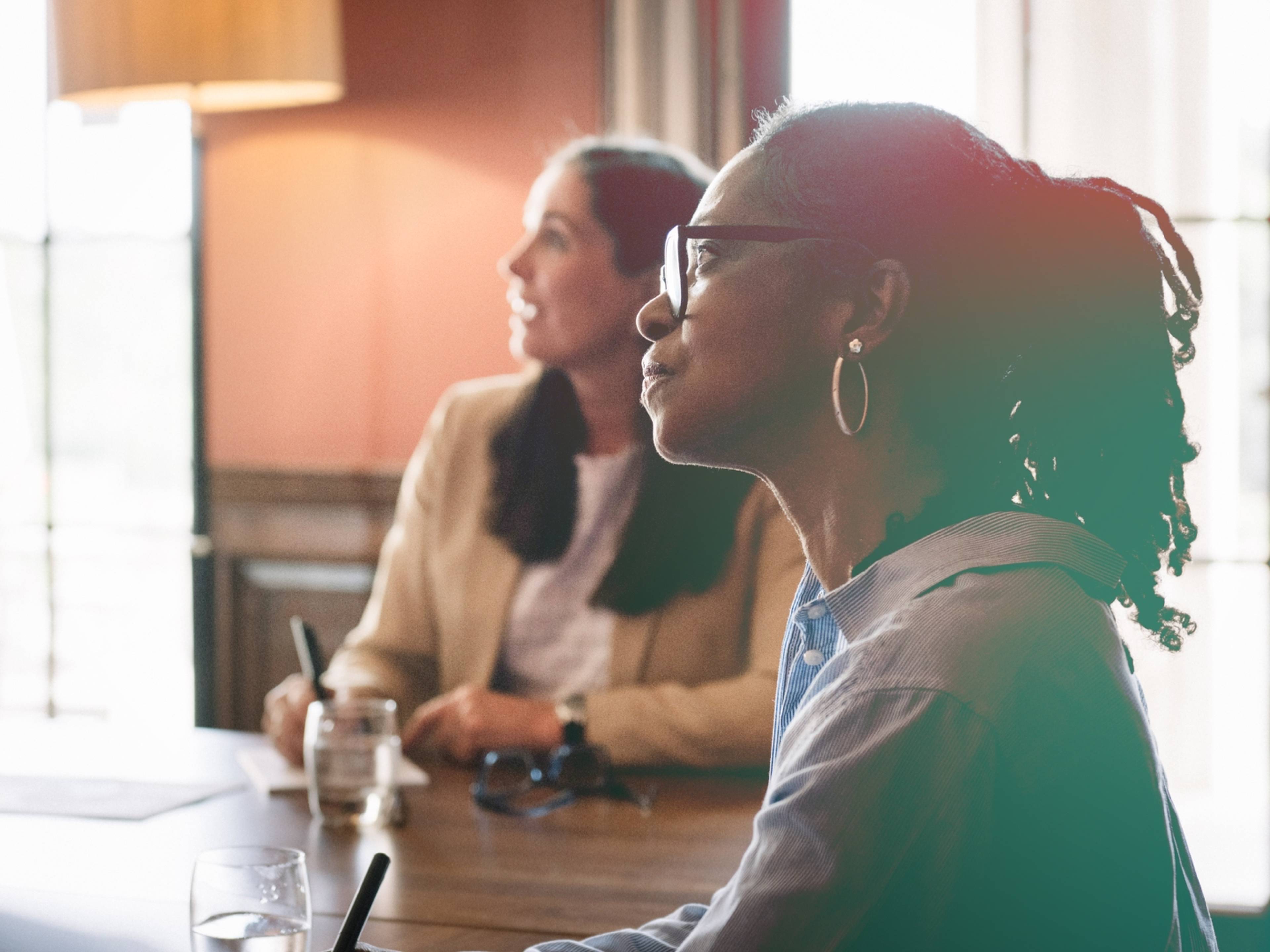 Two women sit at a table, attentively listening during a meeting.