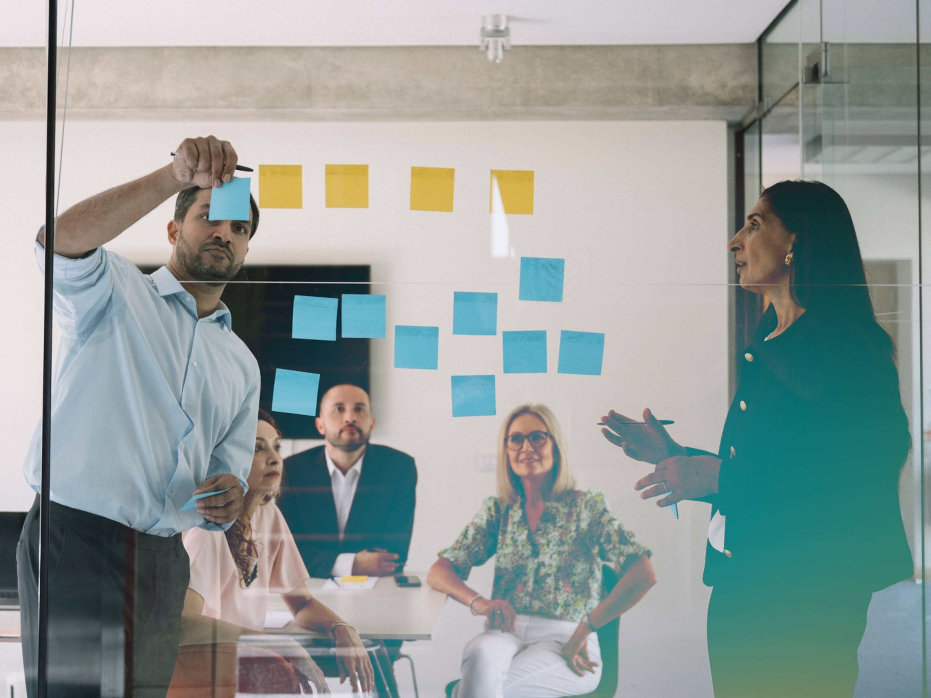 Team in a meeting room, discussing ideas with colorful sticky notes on glass; one person writes while another speaks, three seated listening.