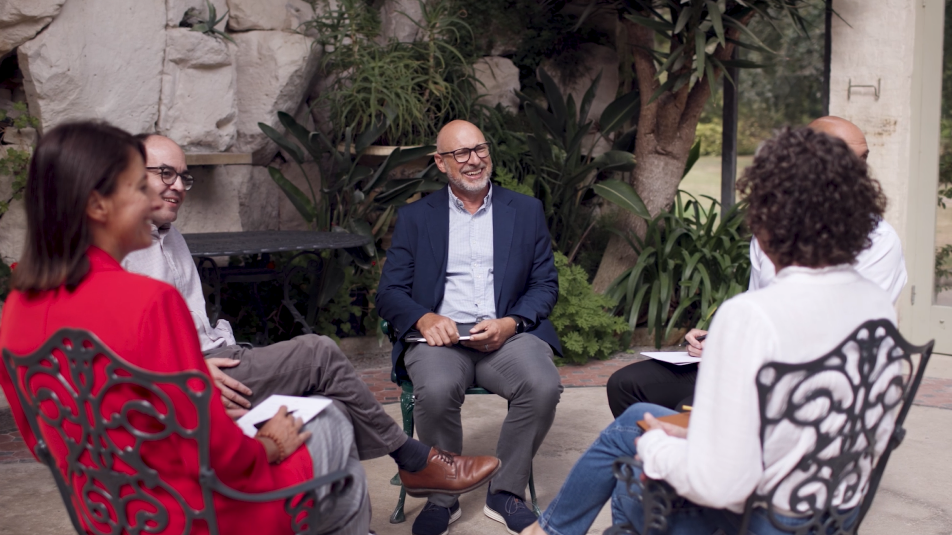 A group of five people sitting in a circle, engaged in a conversation, outdoors near greenery and stone walls.