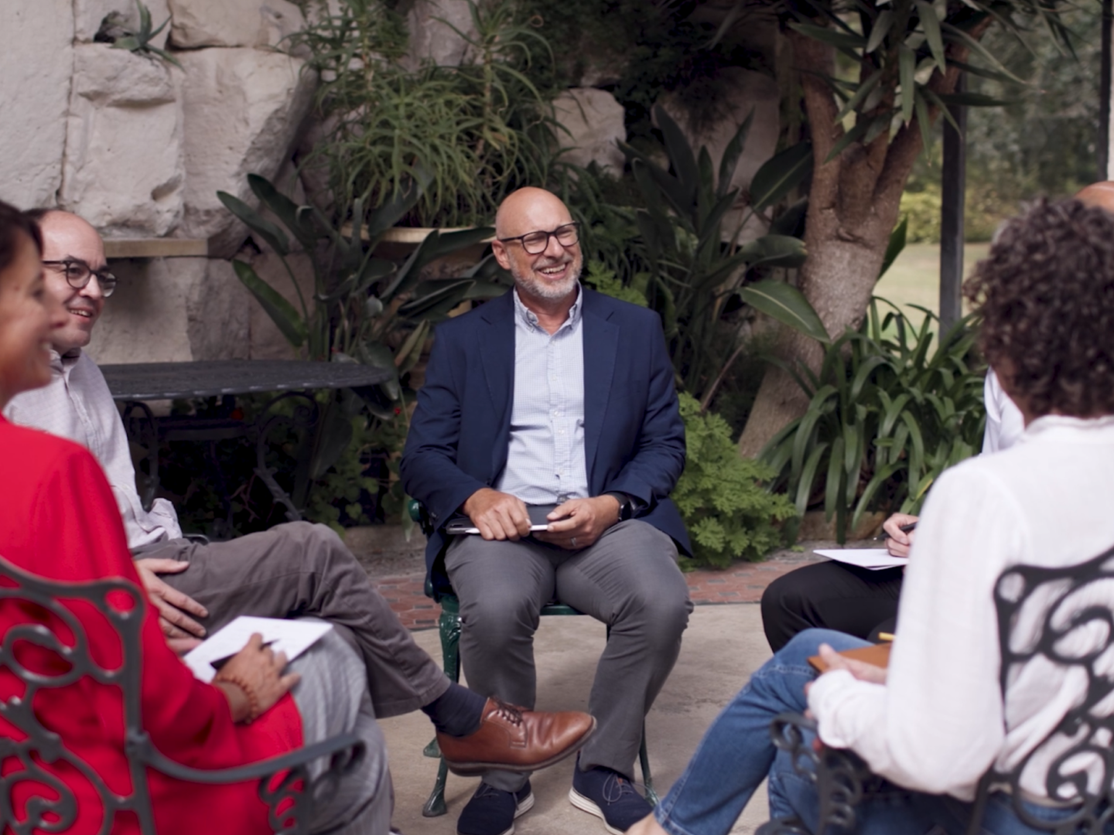 A group of five people sitting in a circle, engaged in a conversation, outdoors near greenery and stone walls.