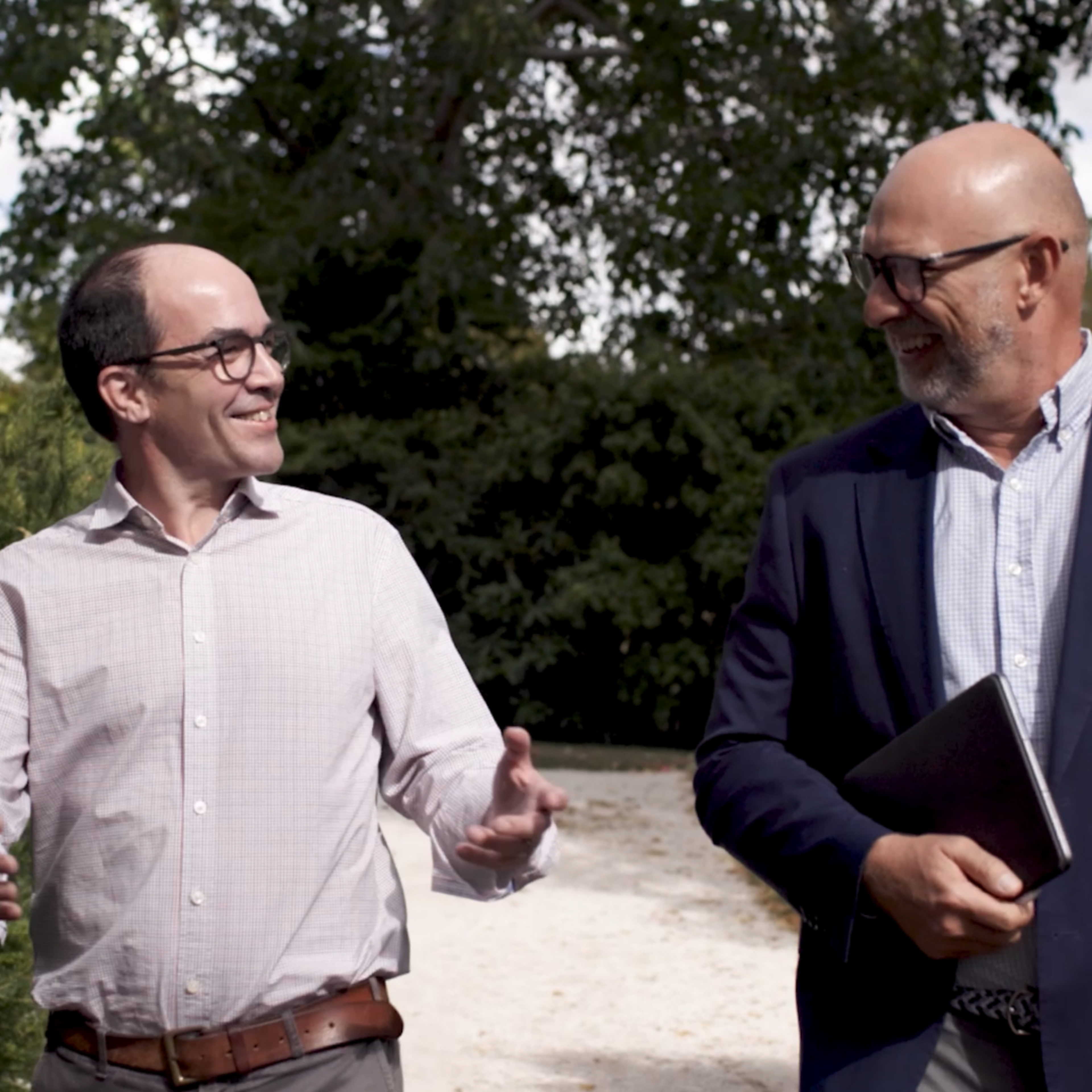 Two men in conversation, walking outdoors. One holds a tablet, both dressed professionally, with trees in the background.