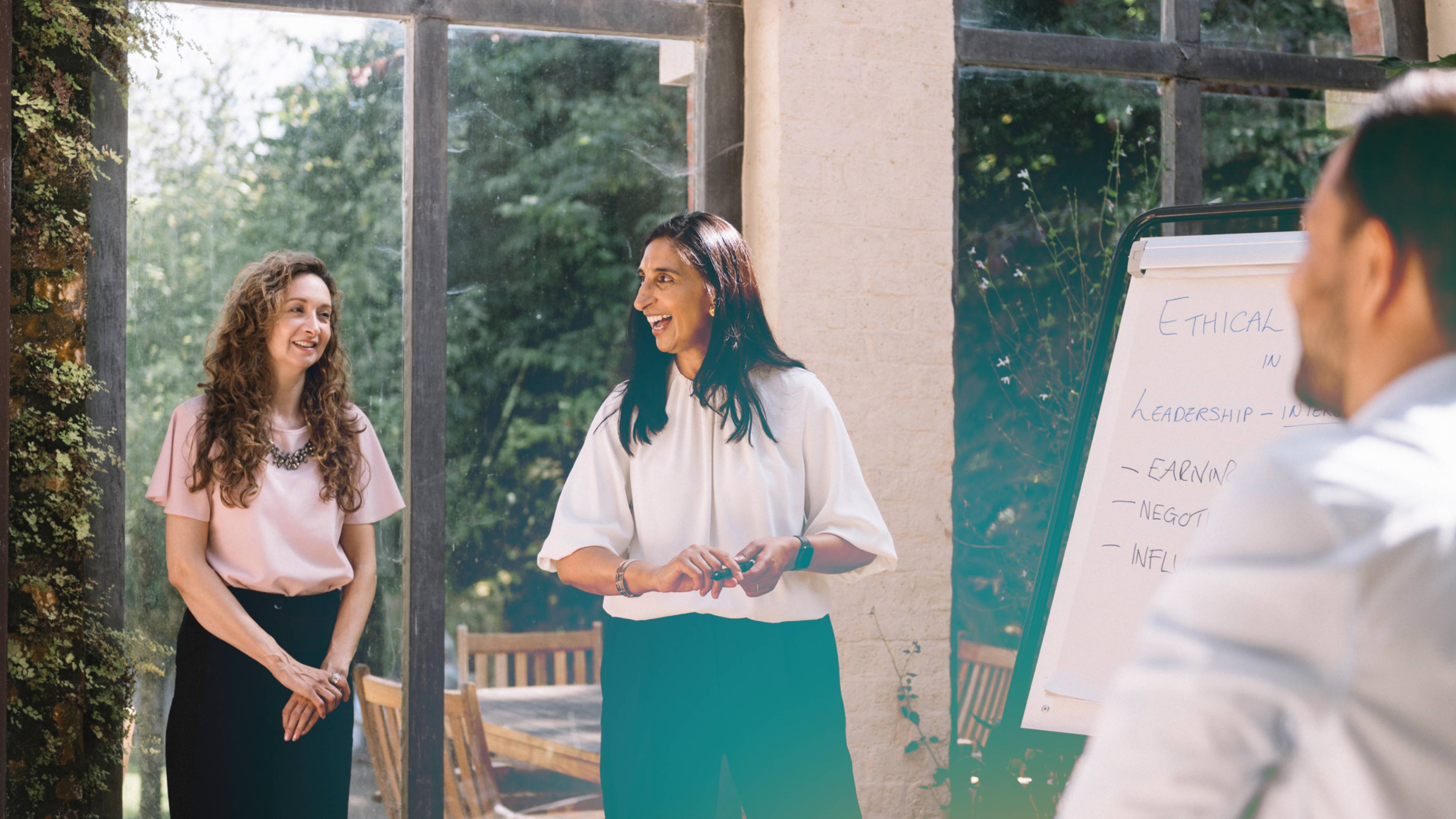 Two leaders stand near a flip chart, smiling and talking, in a bright room with large windows and greenery outside..