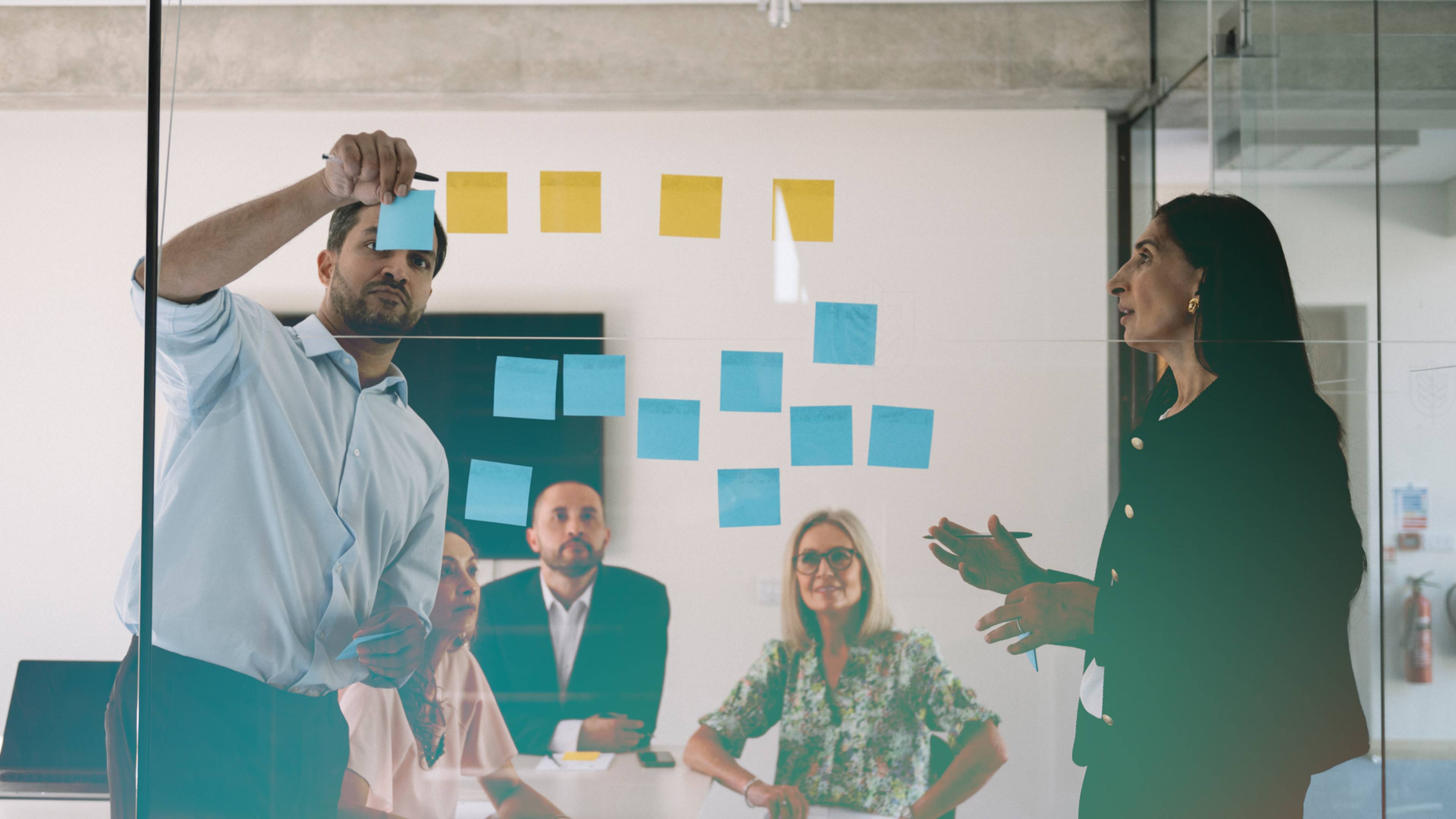 A man places sticky notes on a glass wall, while colleagues seated and standing nearby discuss, in a modern office setting.