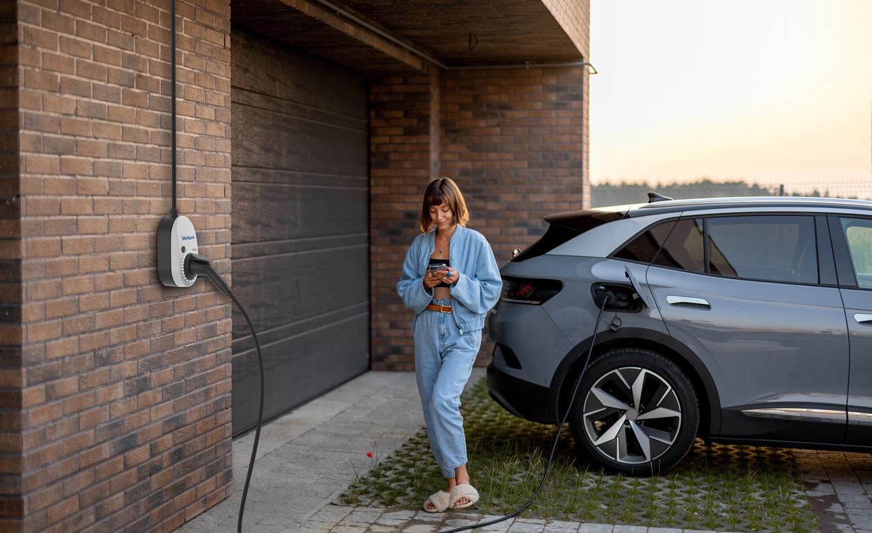 Eine junge Frau steht neben einem E-Auto, lädt dieses und sieht währenddessen auf ihr Smartphone.