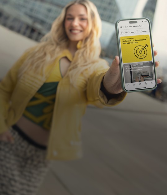 Smiling person holding a smartphone showing a financial app with a yellow target graphic and text in German. The background is an urban setting.