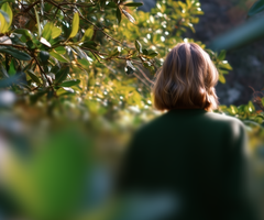 A person with a green coat walks through lush green foliage, seen from behind.