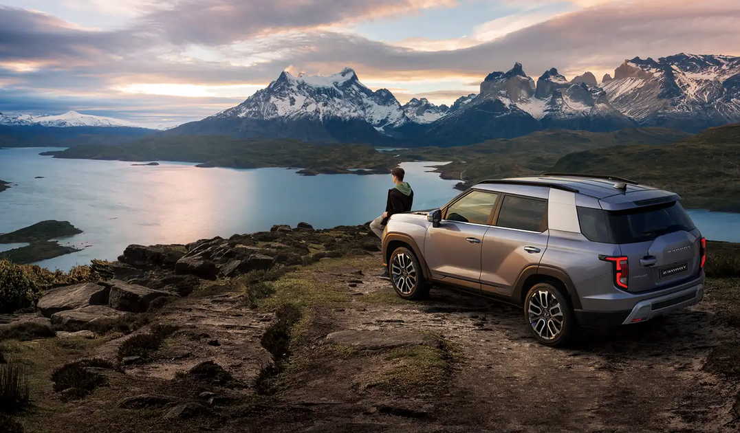 SUV parked on a rocky cliff overlooking a serene lake and snow-capped mountains at sunrise, with a person standing nearby.