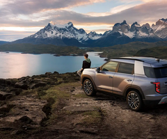 SUV parked on a rocky cliff overlooking a serene lake and snow-capped mountains at sunrise, with a person standing nearby.