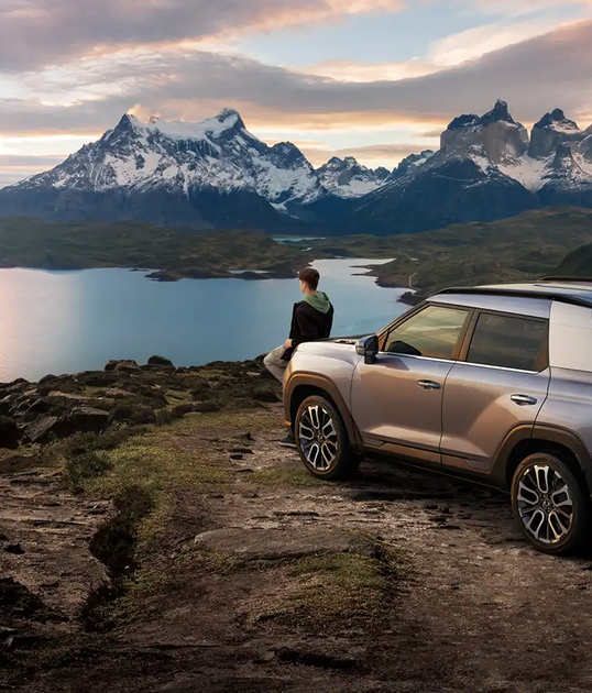 SUV parked on a rocky cliff overlooking a serene lake and snow-capped mountains at sunrise, with a person standing nearby.