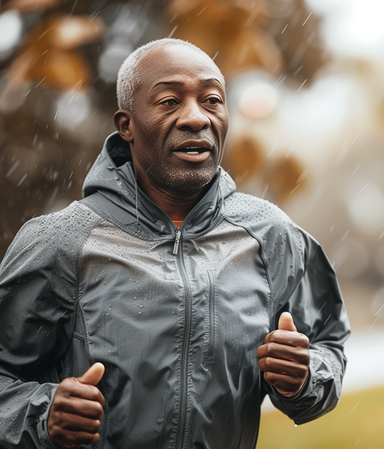 Man jogging in the rain wearing a gray jacket, with blurred autumn trees in the background.