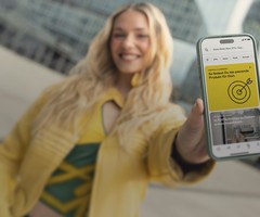 Smiling person holding a smartphone showing a financial app with a yellow target graphic and text in German. The background is an urban setting.