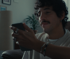 A person with curly hair and a mustache sits on a couch, looking at a smartphone. They wear rings, a bracelet, and a light-colored sweater.