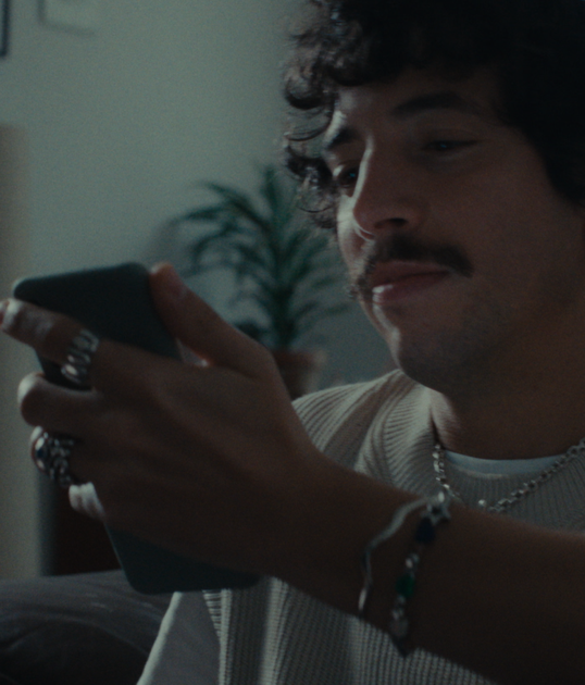 A person with curly hair and a mustache sits on a couch, looking at a smartphone. They wear rings, a bracelet, and a light-colored sweater.