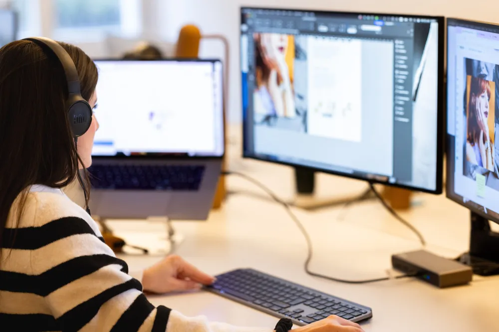 Eine Mitarbeiterin der Valencia trägt Kopfhörer und arbeitet konzentriert an einem Grafikdesign-Projekt. Vor ihr stehen zwei grosse Bildschirme, auf denen eine Designsoftware mit Bildern und Layouts geöffnet ist. Ihr Laptop ist auf einem Ständer platziert, und sie nutzt eine externe Tastatur für effizientes Arbeiten.