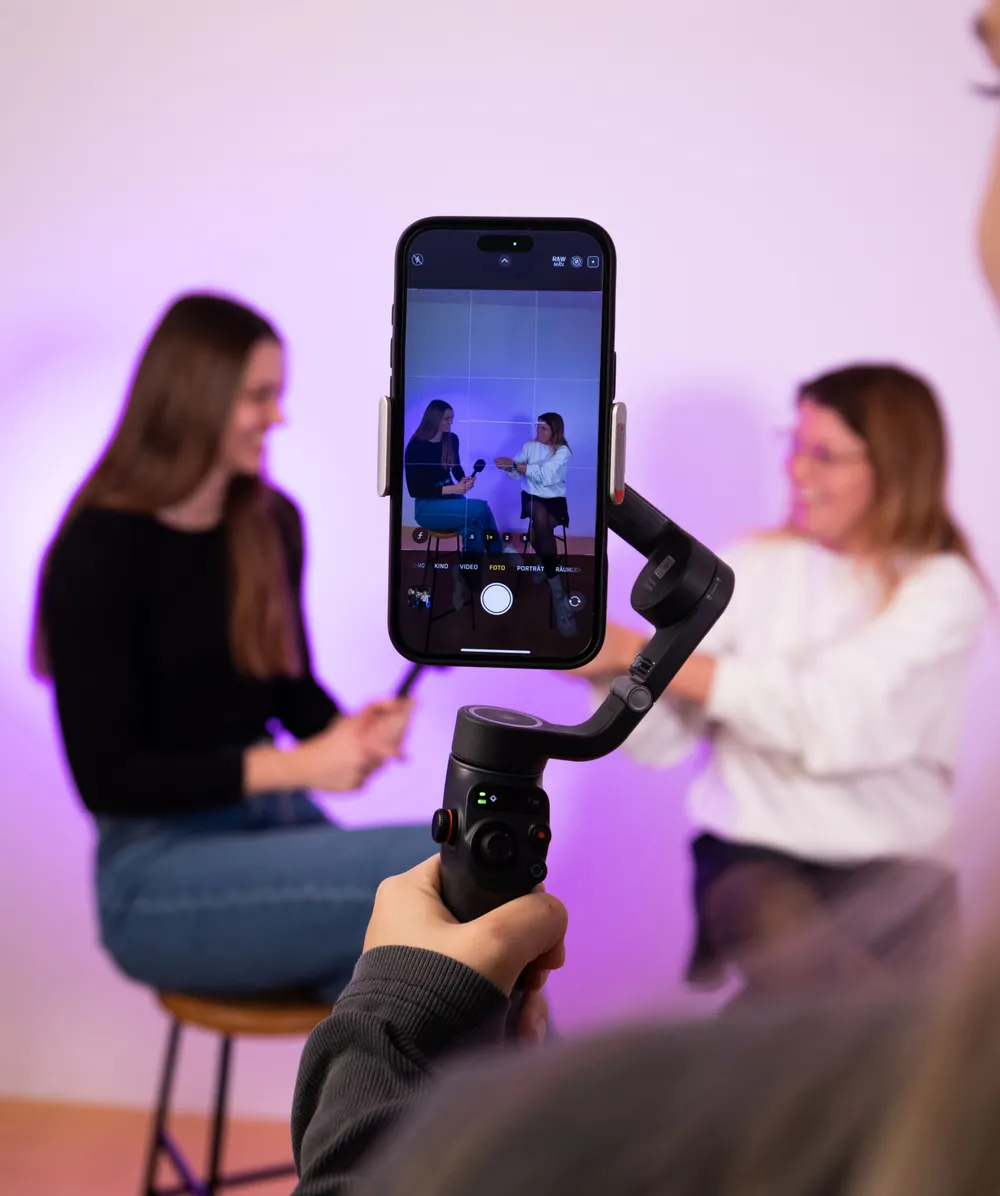 Smartphone auf Gimbal filmt zwei Personen auf Hockern vor lila Hintergrund in Interview-Setting.