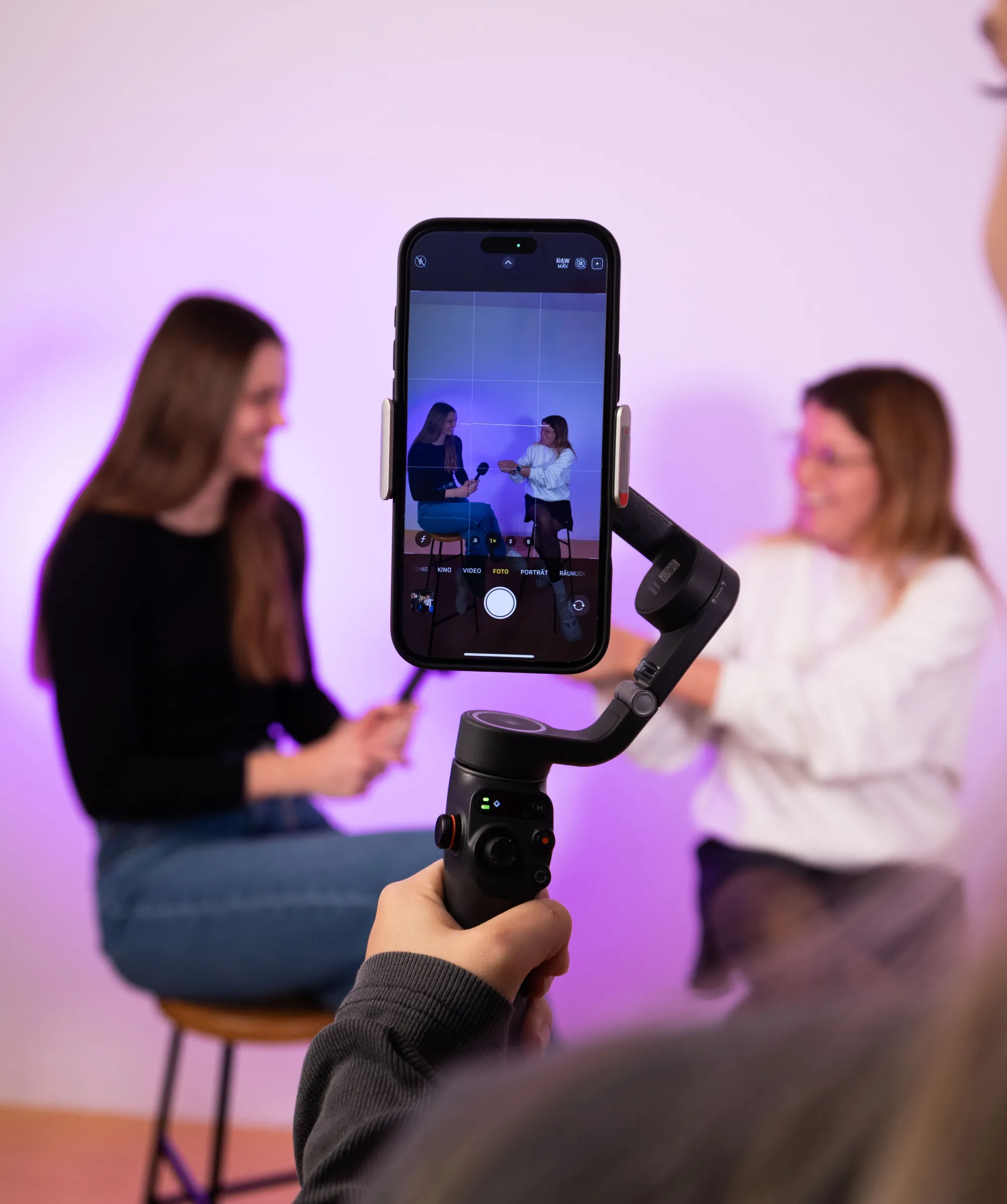 Smartphone auf Gimbal filmt zwei Personen auf Hockern vor lila Hintergrund in Interview-Setting.