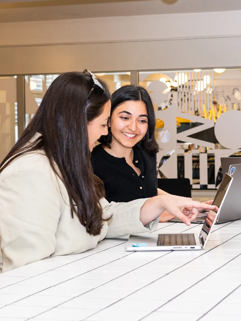 Zwei Frauen sitzen an einem weissen, gefliesten Tisch in einem modernen Büro und schauen gemeinsam auf einen Laptop. Eine von ihnen zeigt lächelnd auf den Bildschirm, während im Hintergrund grafisch gestaltete Fenster die kreative Atmosphäre unterstreichen.