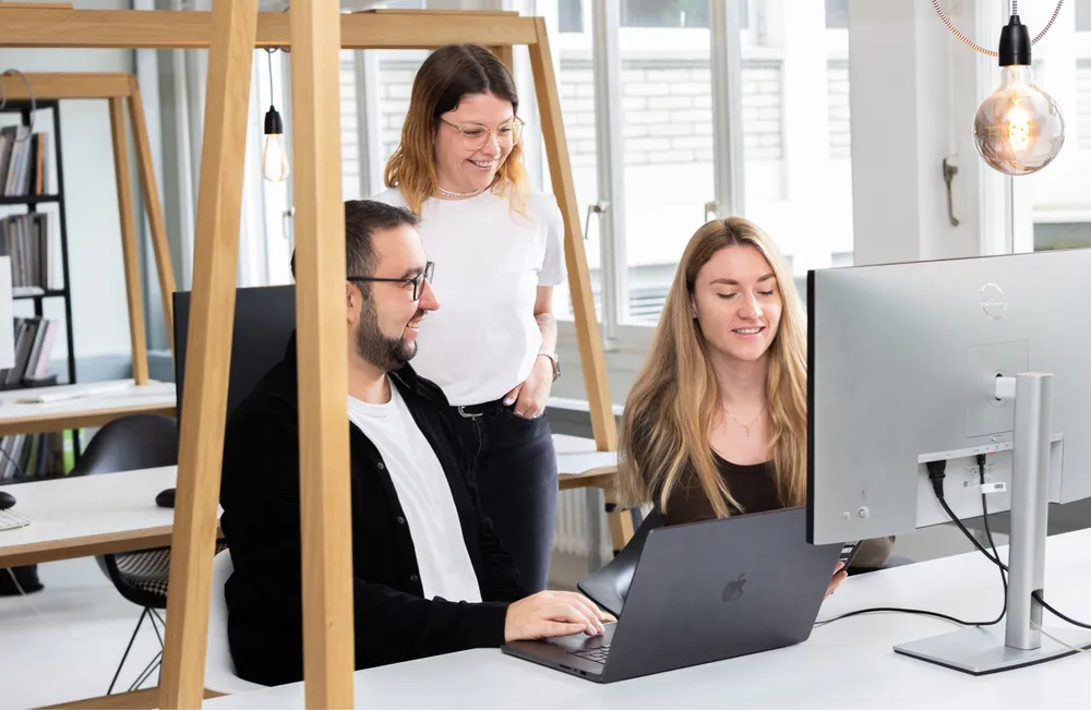 Drei junge Erwachsene arbeiten gemeinsam an einem Schreibtisch mit Monitor und Laptop in einem modernen, hellen Büro. Zwei sitzen nebeneinander und schauen auf den Bildschirm, während eine dritte Person lächelnd daneben steht. Die Stimmung wirkt freundlich und kollegial.