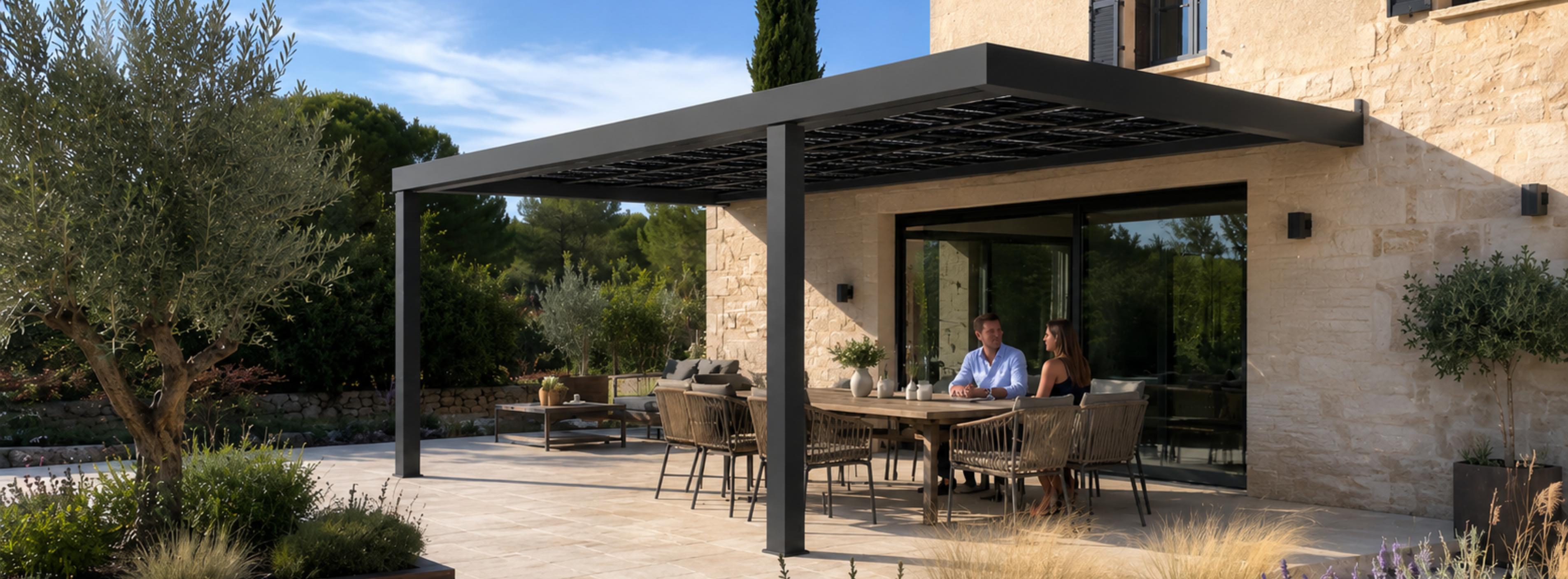 Un couple est assis à une table sous une pergola solaire moderne, sur une terrasse en pierre, à côté d'une maison entourée d'une végétation luxuriante et d'arbres.