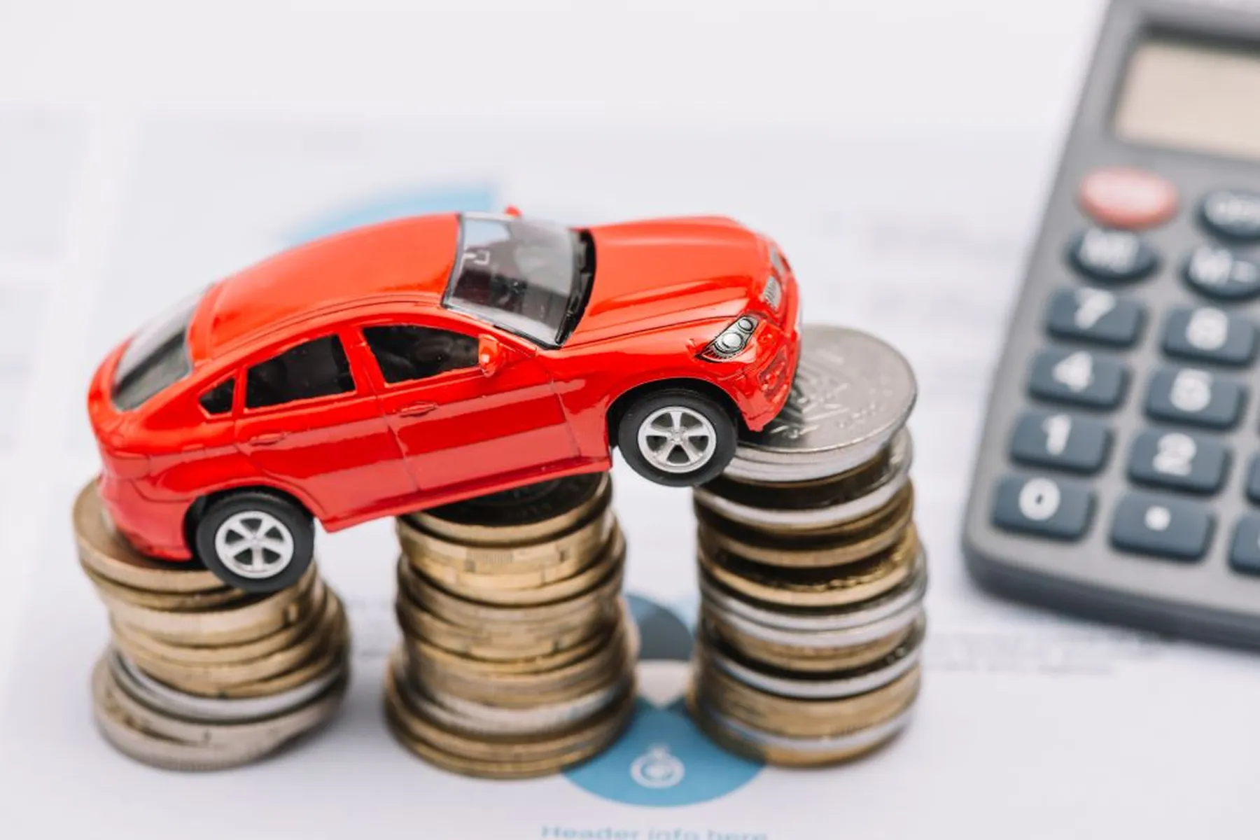Red toy car perched atop stacks of coins with a calculator nearby on paperwork.