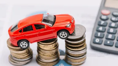 Red toy car perched atop stacks of coins with a calculator nearby on paperwork.