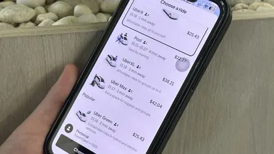 A hand holds a smartphone displaying a ride-sharing app with various ride options and prices, set against a background of smooth white stones.