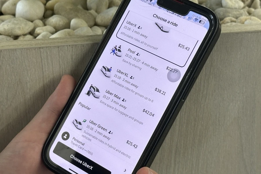 A hand holds a smartphone displaying a ride-sharing app with various ride options and prices, set against a background of smooth white stones.