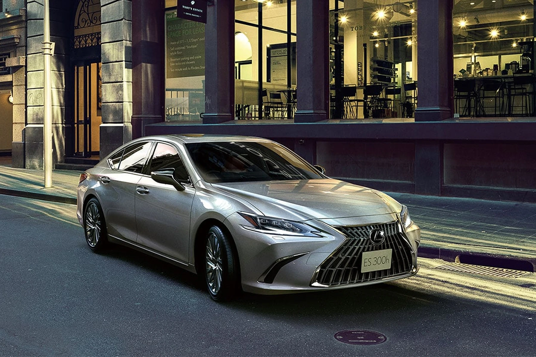 A silver Lexus ES300h parked on a city street at dusk, with lit storefronts in the background.