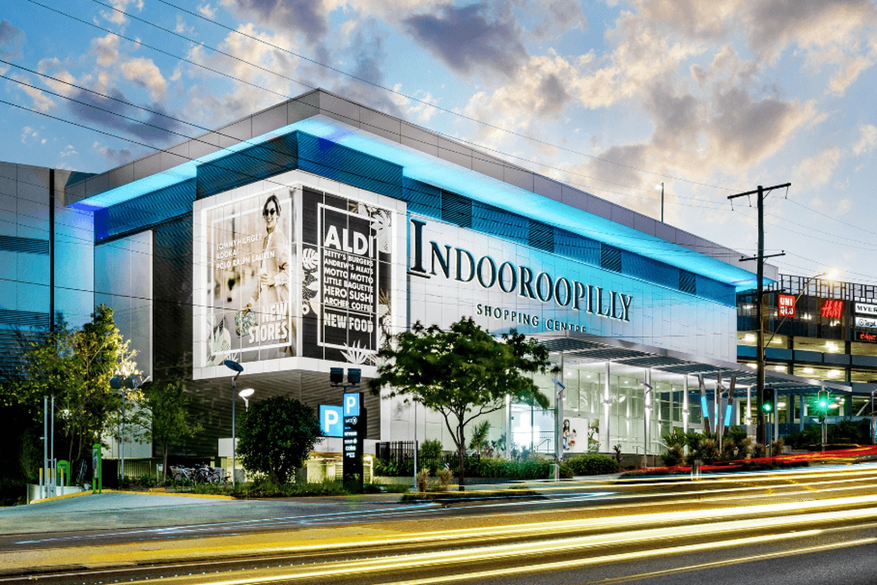 Exterior view of Indooroopilly Shopping Centre at dusk, featuring a modern design, prominent signage, and light trails from passing cars.