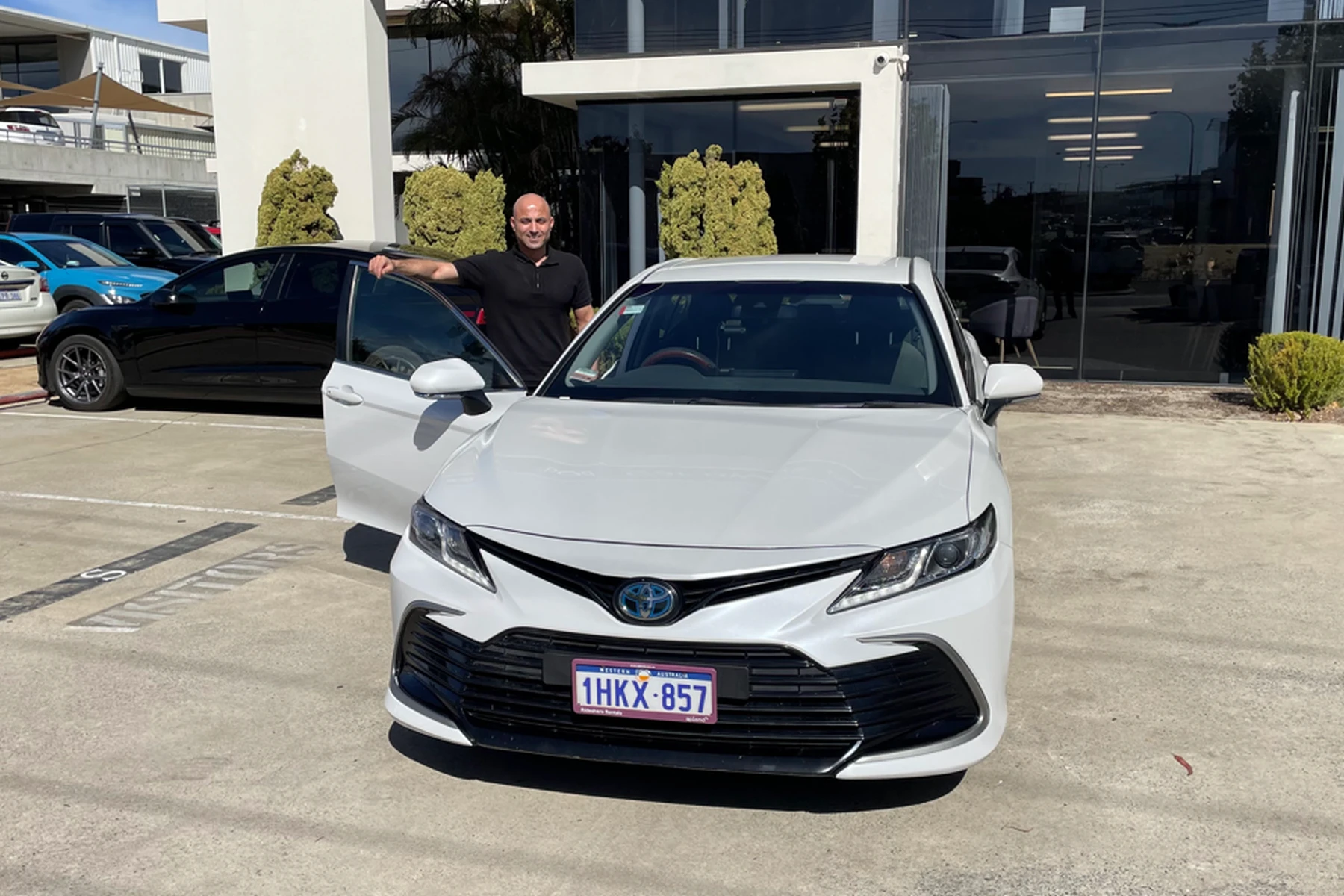 A person stands by a white Toyota sedan in a parking lot, smiling. Other cars and a building are in the background.