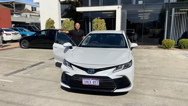A person stands by a white Toyota sedan in a parking lot, smiling. Other cars and a building are in the background.