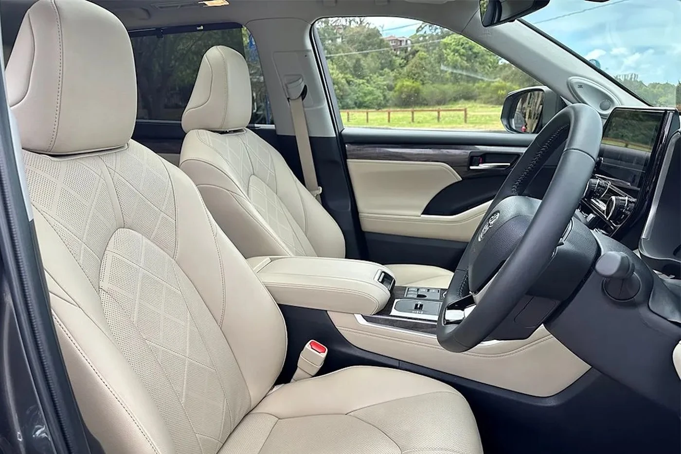 Front interior of a car with light beige leather seats, a black steering wheel, and a view of a grassy landscape through the windows.