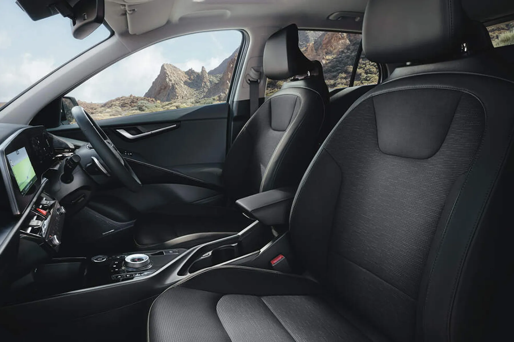 Modern car interior with black leather front seats, dashboard touchscreen, steering wheel and center console; rocky desert seen through windows.