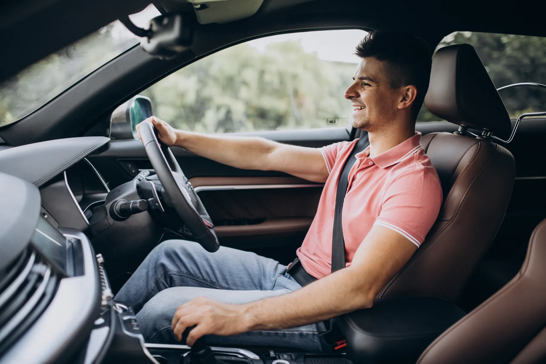 Man in a pink shirt driving a car, smiling, with trees visible through the window.