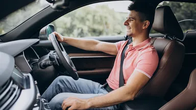 Man in a pink shirt driving a car, smiling, with trees visible through the window.