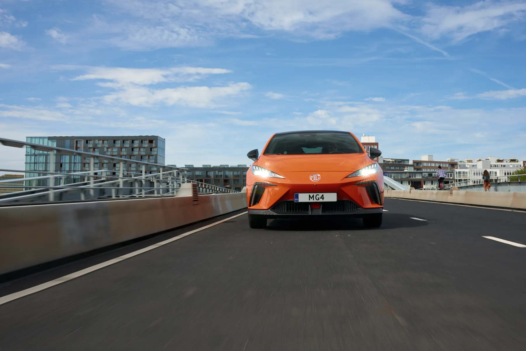 Front-facing orange MG4 hatchback driving on an urban bridge with modern buildings and a blue sky overhead.