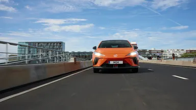 Front-facing orange MG4 hatchback driving on an urban bridge with modern buildings and a blue sky overhead.