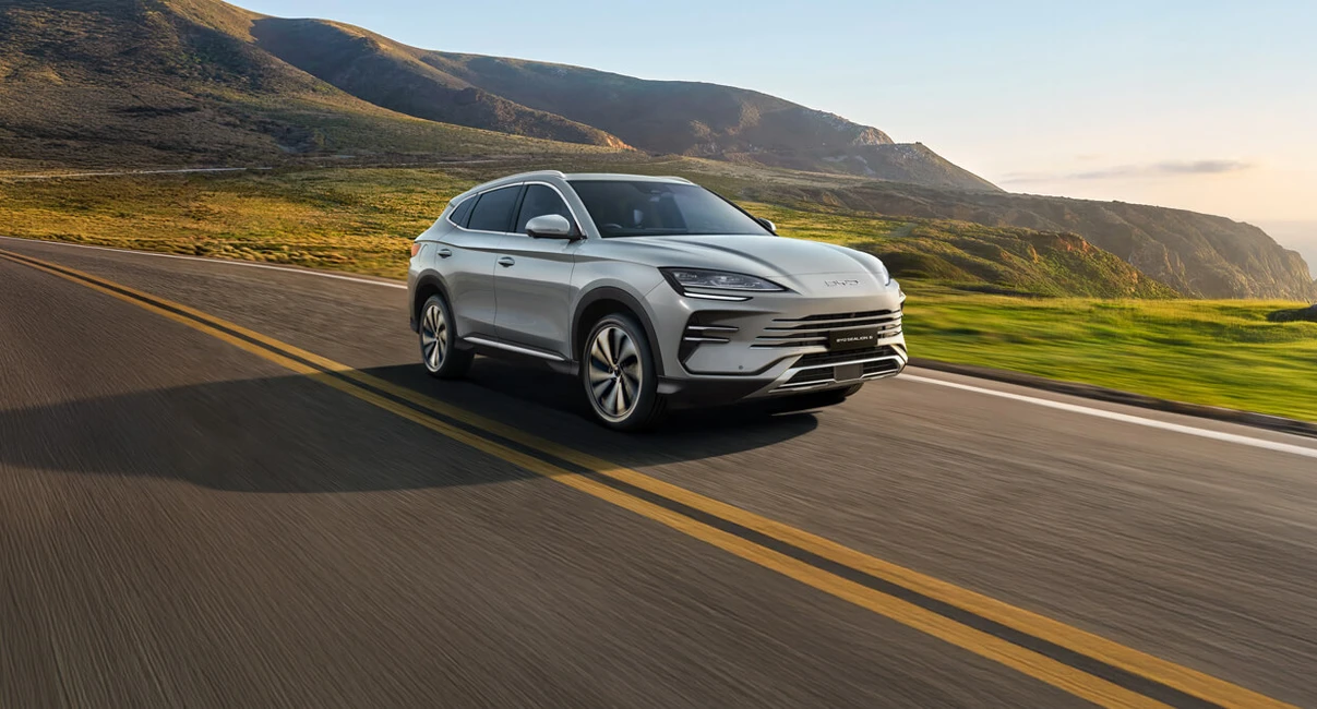 A silver SUV drives on a scenic coastal road with green hills and a clear sky in the background.