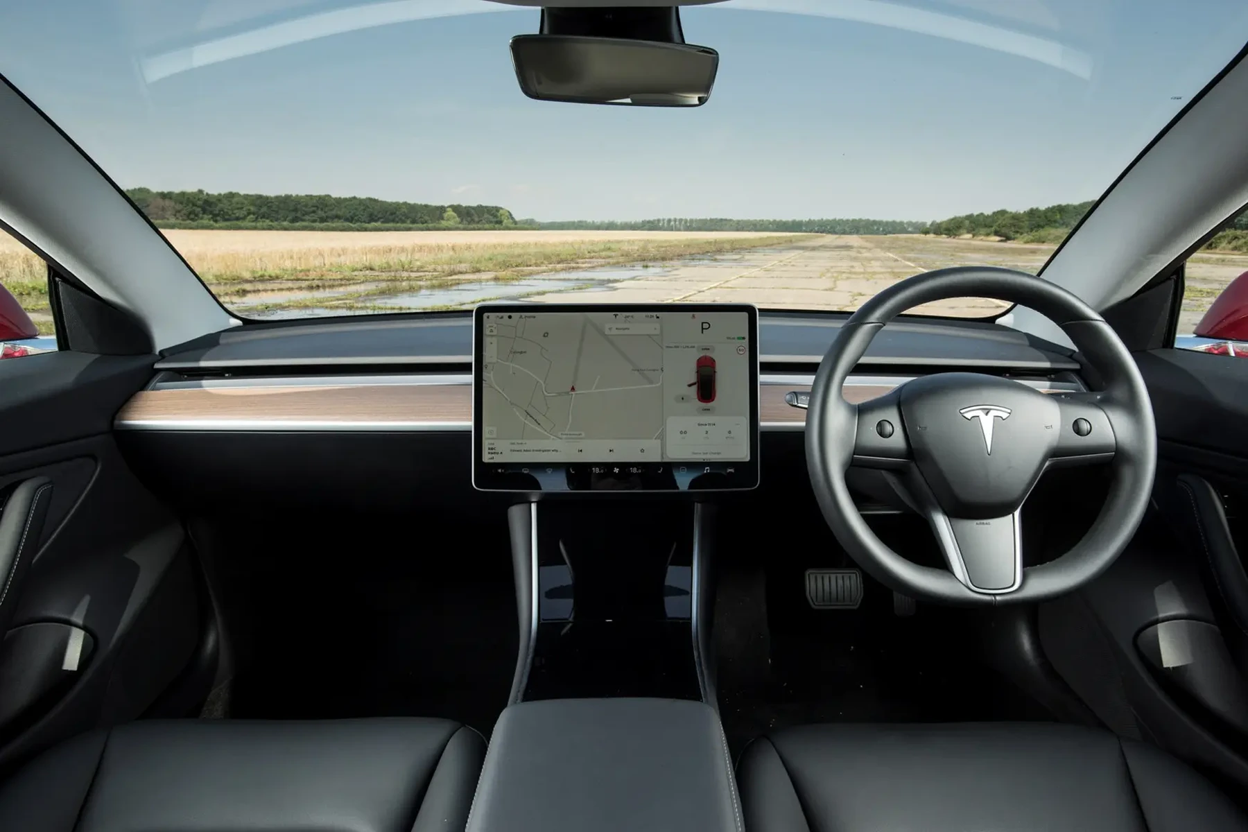 Right-hand-drive Tesla cabin with central touchscreen and minimalist dashboard, view through windshield of an empty road and grassy fields.
