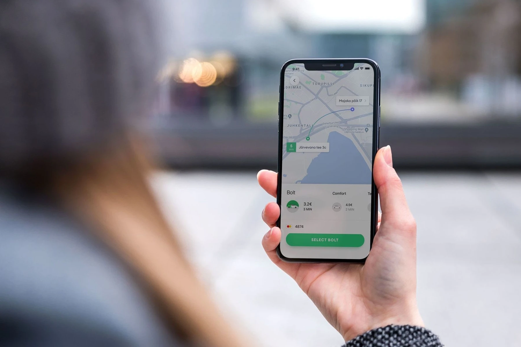 A person holds a smartphone displaying a ride-sharing app with a map and car options, standing outdoors in an urban setting.