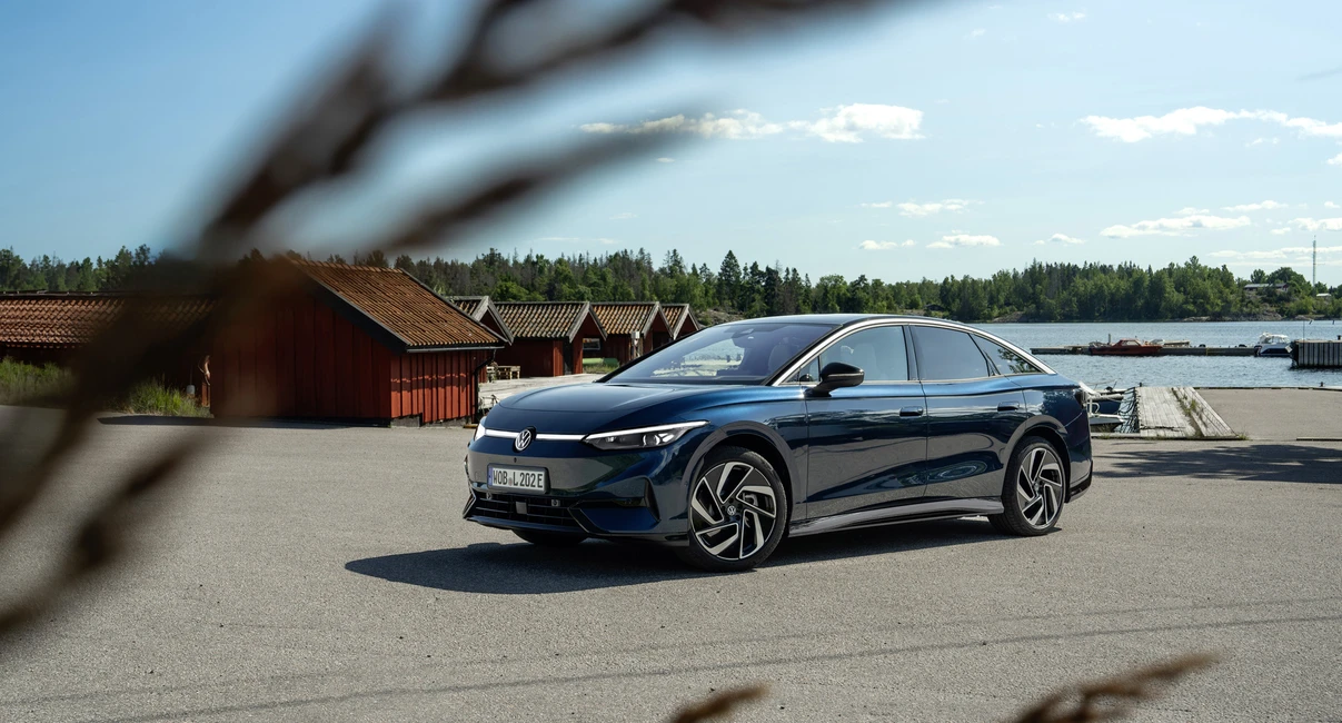 A sleek, dark-colored car parked on a sunny, open area near water, with rustic buildings and trees in the background, framed by blurred grass.
