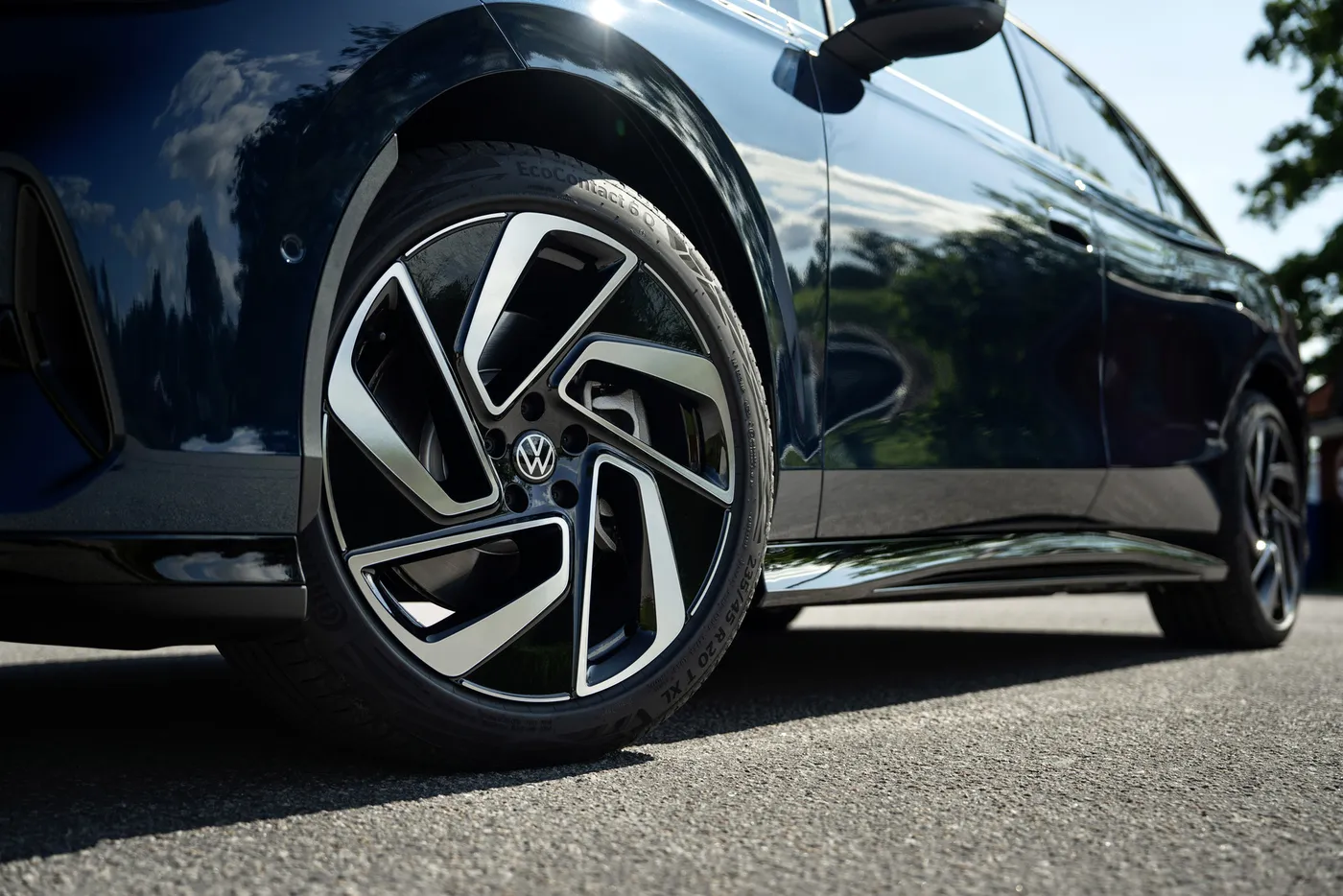 Close-up of a dark blue car with distinctive black and silver alloy wheels, parked on a sunny asphalt road. Reflections visible on car's body.
