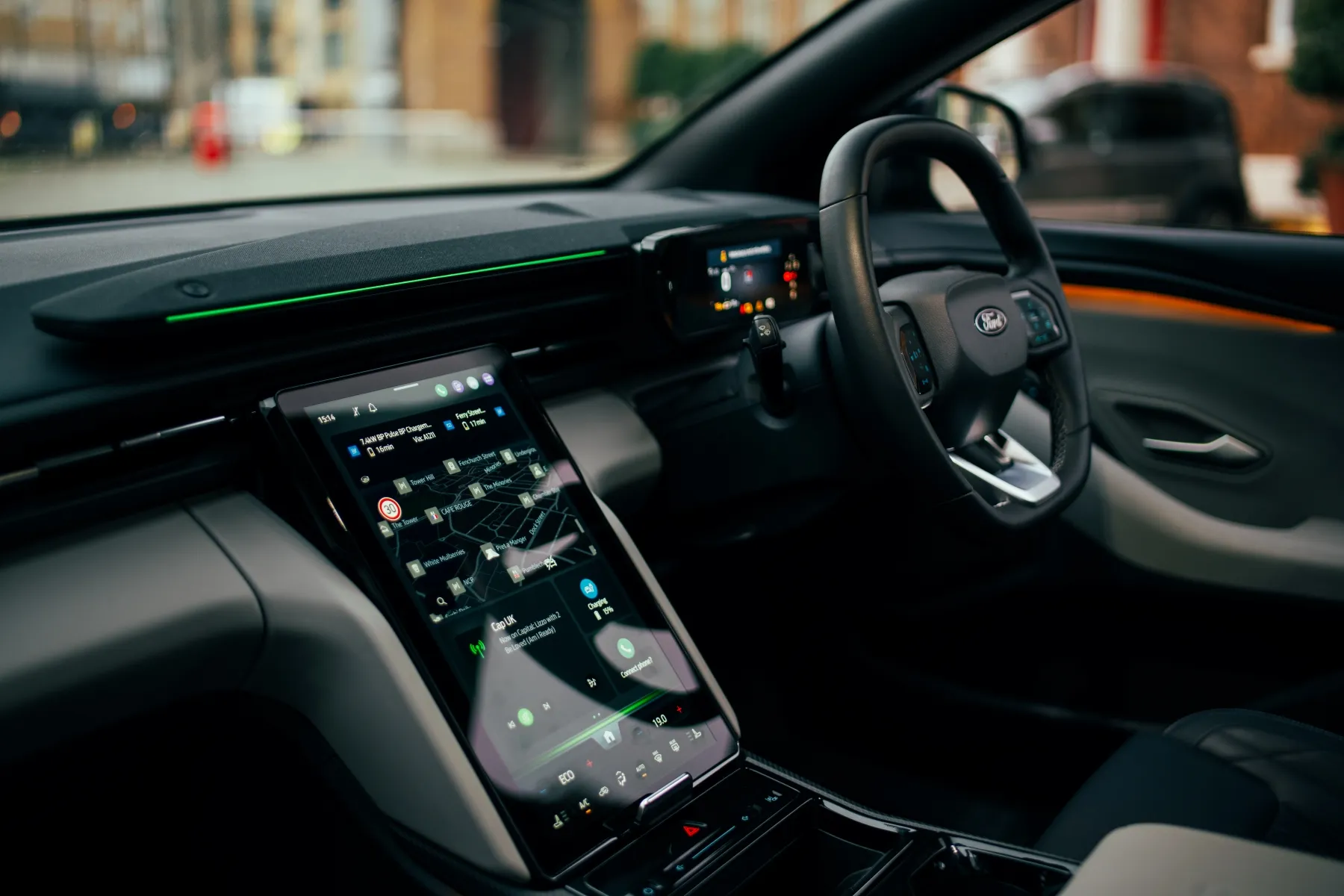 Modern car interior with a large vertical touchscreen center console, digital instrument cluster, ambient green-orange lighting and right-side steering wheel.