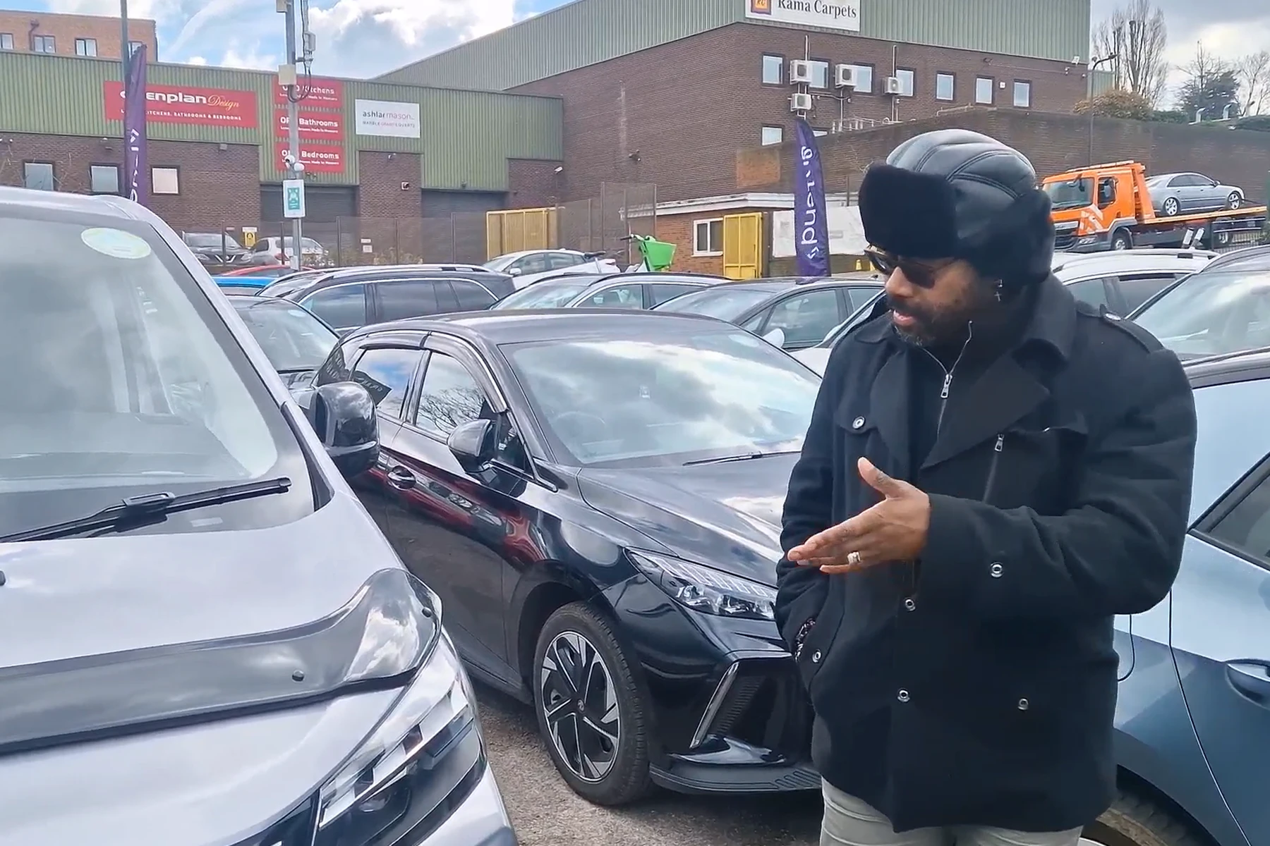 A person in a black coat and hat gestures towards a parked car in a lot filled with various vehicles and industrial buildings in the background.