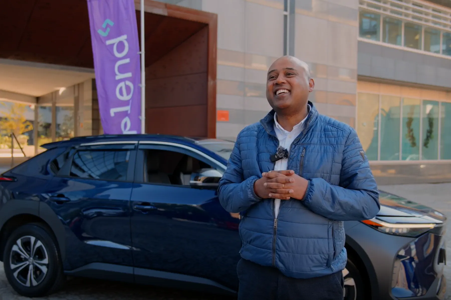 A person in a blue jacket stands smiling beside a parked blue car, with a purple "Lend" banner and modern building in the background.