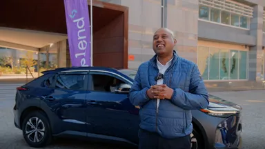 Smiling man in blue jacket holding a microphone standing beside a dark blue SUV outside a modern office building.