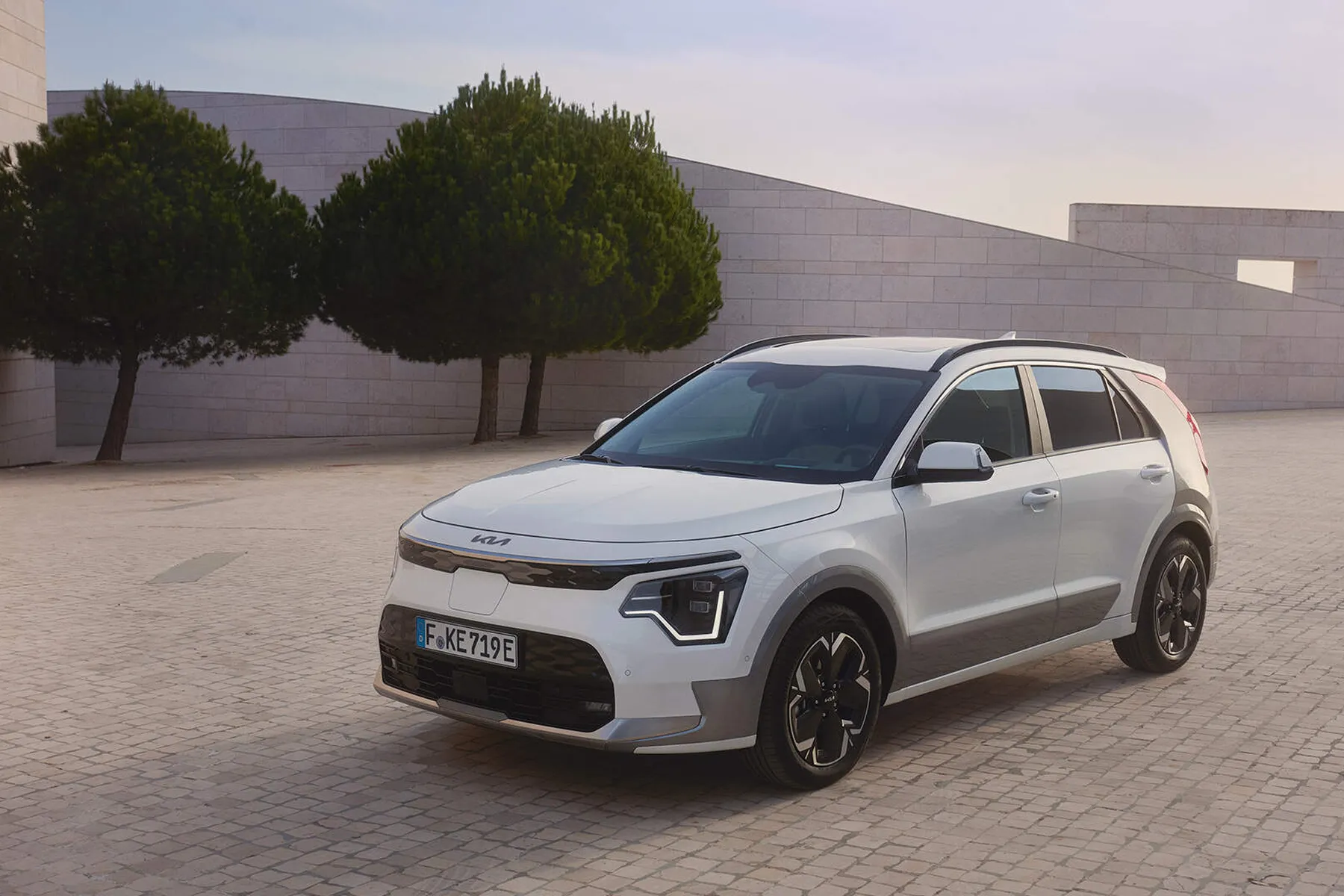 White compact crossover SUV parked on a paved plaza beside modern pale stone walls and two rounded trees under a soft sky.