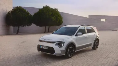 White compact crossover SUV parked on a paved plaza beside modern pale stone walls and two rounded trees under a soft sky.